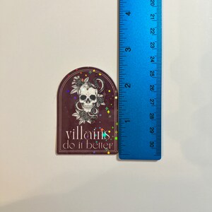 May include: A sticker with a dark red background and a white skull with a snake wrapped around it. The sticker has a holographic finish with rainbow glitter and the text "villains do it better".