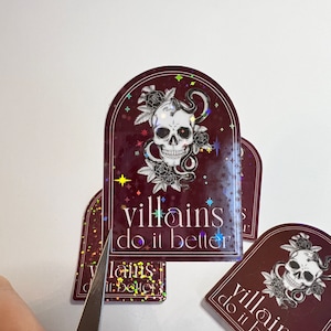 May include: A set of four holographic stickers with a skull and snake design. The stickers are burgundy with gold glitter and the text "villains do it better".