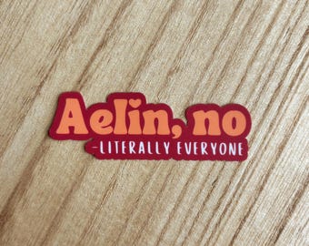 May include: A rectangular sticker with the text "Aelin, no" in orange letters with a heart above the "i", and the text "- LITERALLY EVERYONE" in white letters on a red background. The sticker is on a wooden surface.