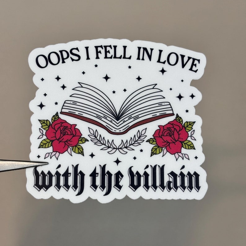 Puede incluir: Pegatina blanca con texto negro que dice "Oops I fell in love with the villain". La pegatina presenta una ilustraci&oacute;n en blanco y negro de un libro abierto con rosas rojas y hojas verdes a cada lado.