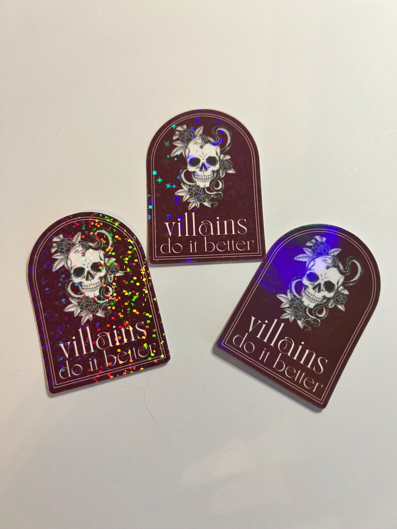 May include: Three holographic stickers with a burgundy background and a skull with a snake wrapped around it. The text on the stickers reads "villains do it better".