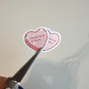 Puede incluir: Dos pegatinas en forma de coraz&oacute;n rosa con el texto "Forbidden Romance" en una y "Forbidden" en la otra. Las pegatinas se est&aacute;n cortando con unas tijeras plateadas.
