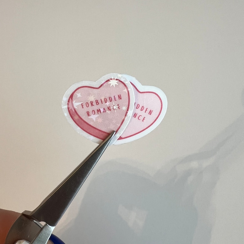 Puede incluir: Dos pegatinas en forma de coraz&oacute;n rosa con el texto "Forbidden Romance" en una y "Forbidden" en la otra. Las pegatinas se est&aacute;n cortando con unas tijeras plateadas.