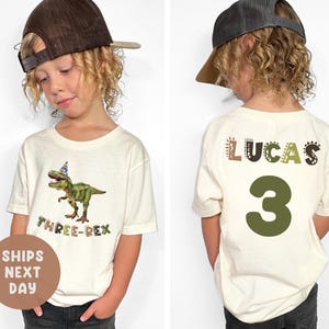 May include: Off-white t-shirt with a green dinosaur wearing a party hat and the text "THREE-REX." The back of the shirt has the name "LUCAS" and the number "3" in green. The child is wearing a brown baseball cap.