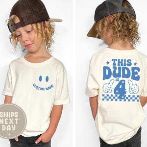 May include: Off-white short-sleeve t-shirt with a blue smiley face and the text "CUSTOM NAME" on the front. The back of the shirt says "THIS DUDE IS 4" in blue with thumbs up and a checkered pattern.