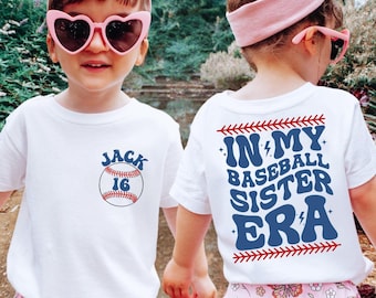 May include: Two children wearing white t-shirts with baseball-themed designs. The child on the left has a baseball with the number 16 and the name "JACK" on their shirt. The child on the right has the text "IN MY BASEBALL SISTER ERA" on their shirt.