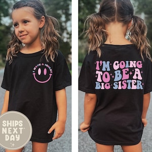 Puede incluir: Camiseta negra con el texto "I HAVE A SECRET" y una carita sonriente rosa en la parte delantera. La parte trasera de la camiseta dice "I'M GOING TO BE A BIG SISTER" en letras rosas, azules y blancas. Una linda camiseta de anuncio.
