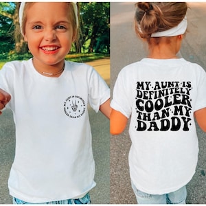 Könnte beinhalten: Weißes T-Shirt mit schwarzem Text, der in einer Retro-Schrift "My aunt is definitely cooler than my daddy" lautet. Der Text befindet sich auf der Rückseite des Shirts. Die Vorderseite des Shirts hat ein kleineres Design mit einem Peace-Zeichen und dem Text "My aunt is definitely cooler than my daddy".