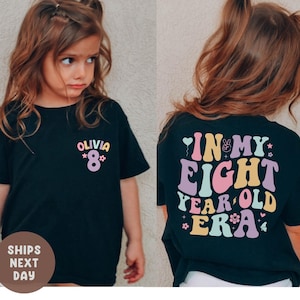 Op de afbeelding: Zwart T-shirt met de tekst "OLIVIA *8*" op de voorkant en "IN MY EIGHT YEAR OLD ERA" op de achterkant. De tekst is in een kleurrijk, retro lettertype. Het shirt wordt gedragen door een jong meisje met bruin haar.