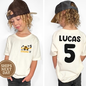 May include: A white t-shirt with a yellow construction truck graphic and the text "I'm 5 and digging it". The back of the shirt has the name "Lucas" and the number "5" in black.