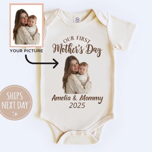 Könnte beinhalten: Ein weißer Baby-Body mit dem Text "Our First Mother's Day" in braunen Buchstaben. Der Body zeigt außerdem ein Foto einer Mutter und ihres Kindes, mit dem Text "Amelia & Mommy 2025" unter dem Foto.