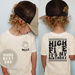 May include: A cream-colored t-shirt with the text "HIGH FIVE IT'S MY BIRTHDAY" on the back and a hand graphic. The front of the shirt has a hand graphic and the name "MILES". The child is wearing a brown and tan trucker hat.
