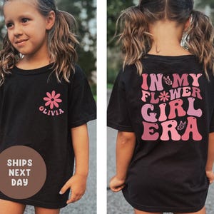 以下が含まれることがあります： ピンクの花のグラフィックと「Olivia」という名前が入った黒いTシャツ。Tシャツの背面には、ピンクと紫の色合いで「In My Flower Girl Era」という文字がプリントされています。カジュアルで快適なTシャツ。
