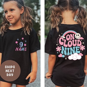 May include: Black t-shirt with a front design featuring the number "9" and the word "NAME" in pink and blue. The back of the shirt has a design that says "ON CLOUD NINE" with a rainbow, flowers, and a smiling face.