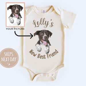 May include: A cream-colored baby onesie with the text "Lolly's New Best Friend" in brown text. A brown and white dog is peeking out of the onesie, with its tongue sticking out.