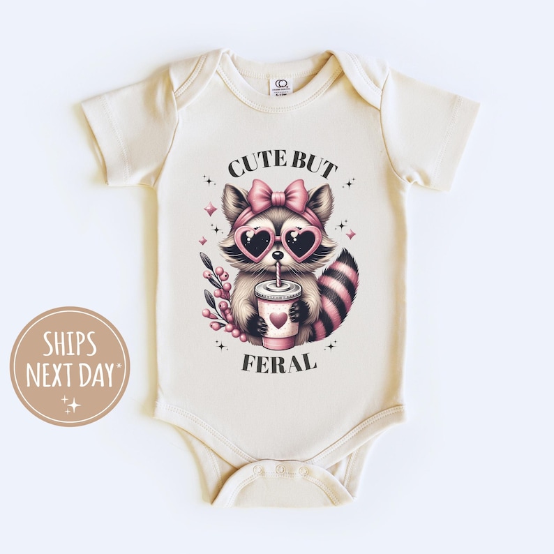 Pu&ograve; includere: Body per neonati color crema con grafica di procione con occhiali da sole a forma di cuore e fiocco. Il procione tiene in mano una bevanda con un cuore. Il testo recita "CUTE BUT FERAL".