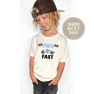 May include: A cream-colored t-shirt featuring a light blue race car design with the text "TWO FAST". The shirt is worn by a young child wearing a brown baseball cap. The shirt is a birthday-themed design.