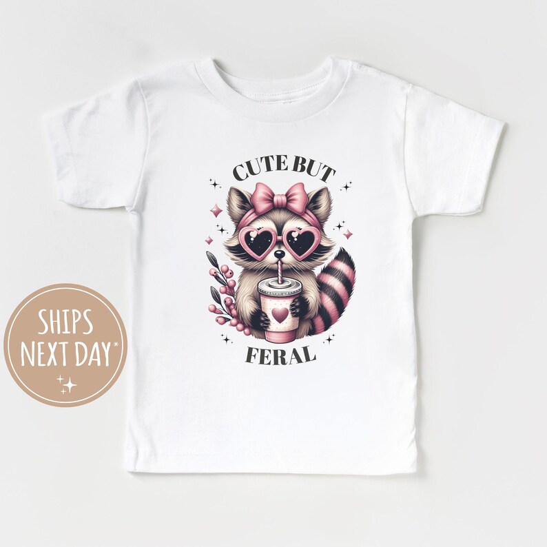 Pu&ograve; includere: T-shirt bianco con un'illustrazione di un procione con occhiali da sole a forma di cuore e un fiocco rosa. Il procione tiene in mano una tazza con un cuore. Il testo "CUTE BUT FERAL" &egrave; stampato sulla maglietta.