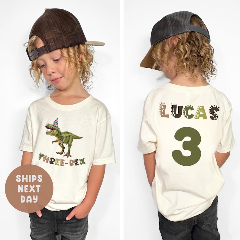 May include: Cream-coloured t-shirt featuring a green dinosaur with a party hat and the text "THREE-REX." The back of the shirt displays the name "LUCAS" and the number "3" in green. The child is wearing a brown baseball cap.