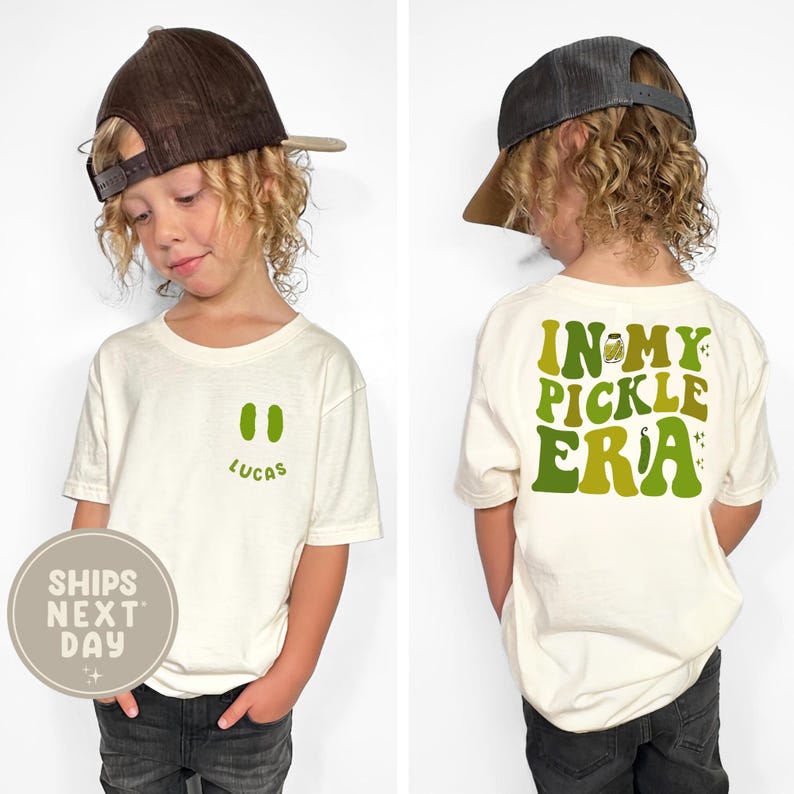May include: Off-white t-shirt with a green pickle design and the text "Lucas". The back of the shirt says "In My Pickle Era" in green lettering. The child is wearing a brown baseball cap and dark jeans.