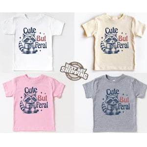 May include: Four toddler t-shirts with a cute raccoon graphic and the text "Cute But Feral". The shirts are in white, beige, pink, and gray.