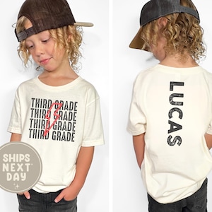 May include: Cream-colored t-shirt with "THIRD GRADE" repeated and crossed out with a red lightning bolt design. The back of the shirt has the name "LUCAS" printed vertically. The child is wearing a brown baseball cap.