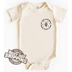 May include: A cream-coloured baby onesie with a black graphic that reads "My aunt is definitely cooler than my daddy" with a peace sign and stars.