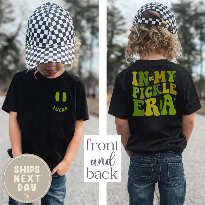 May include: Black t-shirt with the text "IN MY PICKLE ERA" on the back and two green pickle shapes and the name "LUCAS" on the front. The child is wearing a black and white checkered baseball cap and blue jeans.