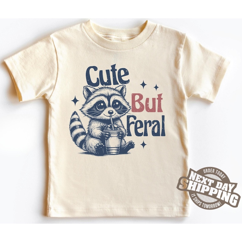 May include: A cream colored t-shirt with a cartoon raccoon drinking a beverage. The raccoon has a cute face and is wearing a blue bandana. The text "Cute But Feral" is printed on the shirt in red and blue.