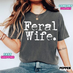 May include: A gray t-shirt with the text "Feral Wife." printed in white. The shirt has a relaxed fit and a crew neck.