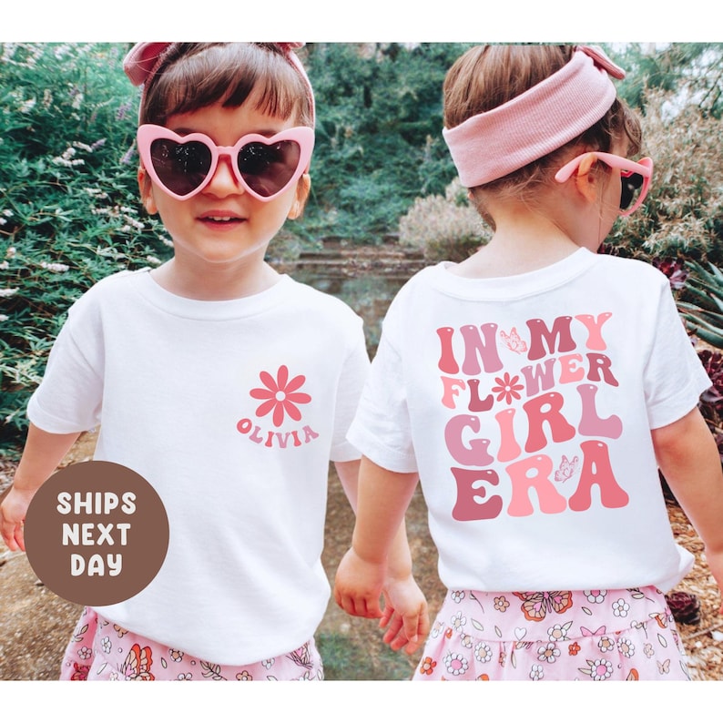 以下が含まれることがあります： 白いTシャツを着た2人の子供たち。花柄のデザインと「Olivia」と「In My Flower Girl Era」の文字がプリントされています。子供たちはピンクのサングラスとヘアバンドも着用しています。シャツは花柄のスカートと組み合わせています。