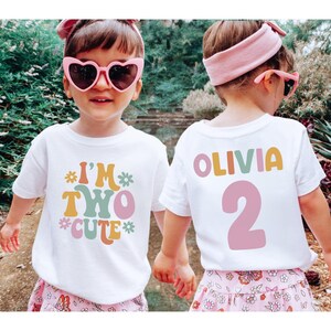 Może przedstawiać: Dwie białe koszulki z retro wzorem. Jedna koszulka ma napis "I'm Two Cute" z kwiatami, a druga "Olivia 2".