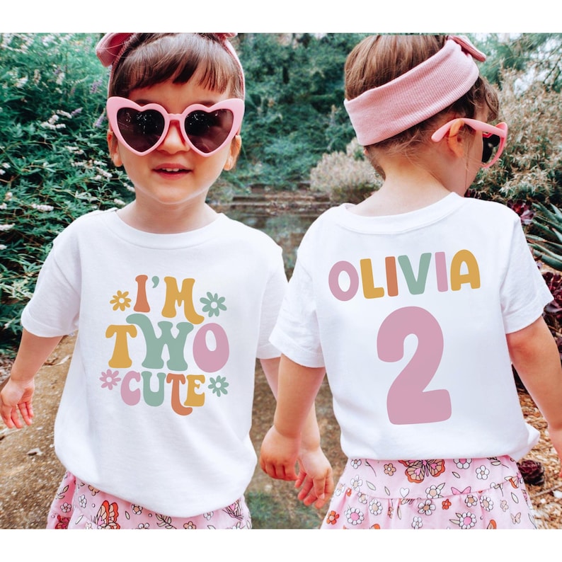Może przedstawiać: Dwie białe koszulki z retro wzorem. Jedna koszulka ma napis "I'm Two Cute" z kwiatami, a druga "Olivia 2".