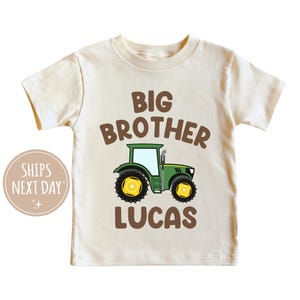 May include: A beige short-sleeved t-shirt with the text "BIG BROTHER" in brown letters above a green and yellow cartoon tractor. The name "LUCAS" is printed in brown letters below the tractor.