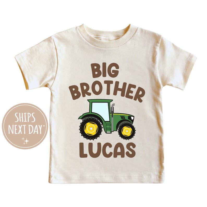 May include: A beige short-sleeved t-shirt with the text "BIG BROTHER" in brown letters above a green and yellow cartoon tractor. The name "LUCAS" is printed in brown letters below the tractor.