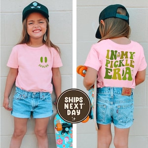 May include: A young person wearing a pink t-shirt with a green pickle graphic and the text "Skylar" on the front. The back of the shirt says "In My Pickle Era" in green. They are wearing blue denim shorts and a black baseball cap. A skateboard is in the image.