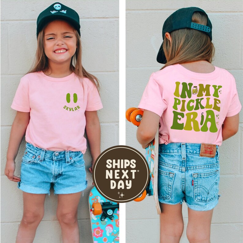 May include: A young person wearing a pink t-shirt with a green pickle graphic and the text "Skylar" on the front. The back of the shirt says "In My Pickle Era" in green. They are wearing blue denim shorts and a black baseball cap. A skateboard is in the image.