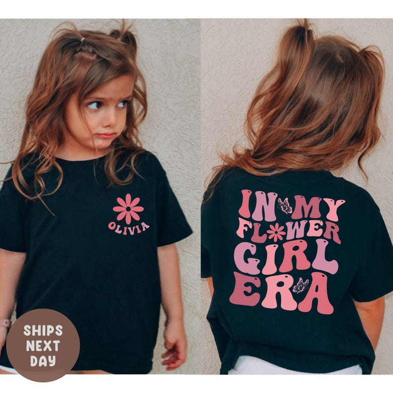 以下が含まれることがあります： 前面にピンクの花と「Olivia」の名前がプリントされた黒いTシャツ。Tシャツの背面には、ピンク色の文字で「In My Flower Girl Era」と書かれています。Tシャツは、おさげ髪の若い女の子が着ています。