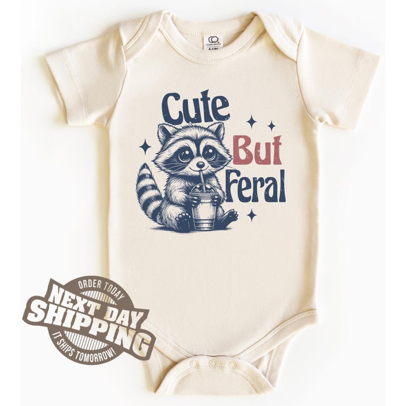 May include: A cream-colored baby onesie with a cartoon raccoon drinking from a cup. The text "Cute But Feral" is printed on the onesie.