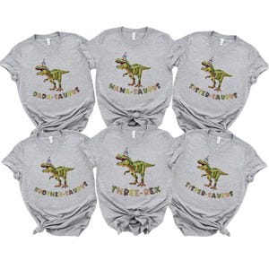 May include: Six grey t-shirts with dinosaur graphics and text. Each shirt features a green dinosaur wearing a party hat. Text includes "DADA-SAURUS", "MAMA-SAURUS", "SISTER-SAURUS", "BROTHER-SAURUS", and "THREE-REX".