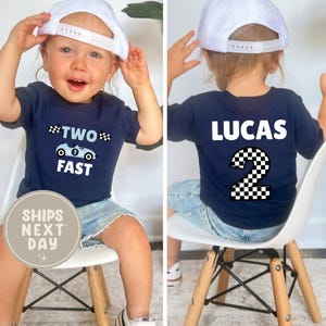 May include: Navy blue toddler t-shirt with the words "TWO FAST" and a race car graphic on the front. The back of the shirt has the name "LUCAS" and a checkered number 2. The child is wearing a white baseball cap.