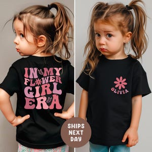 以下が含まれることがあります： ピンクのグラフィックが入った黒いTシャツが2枚。1枚のシャツの背面には「IN MY FLOWER GIRL ERA」と書かれています。もう1枚のシャツにはピンクの花と「OLIVIA」という名前が入っています。どちらのシャツも幼い子供が着ています。