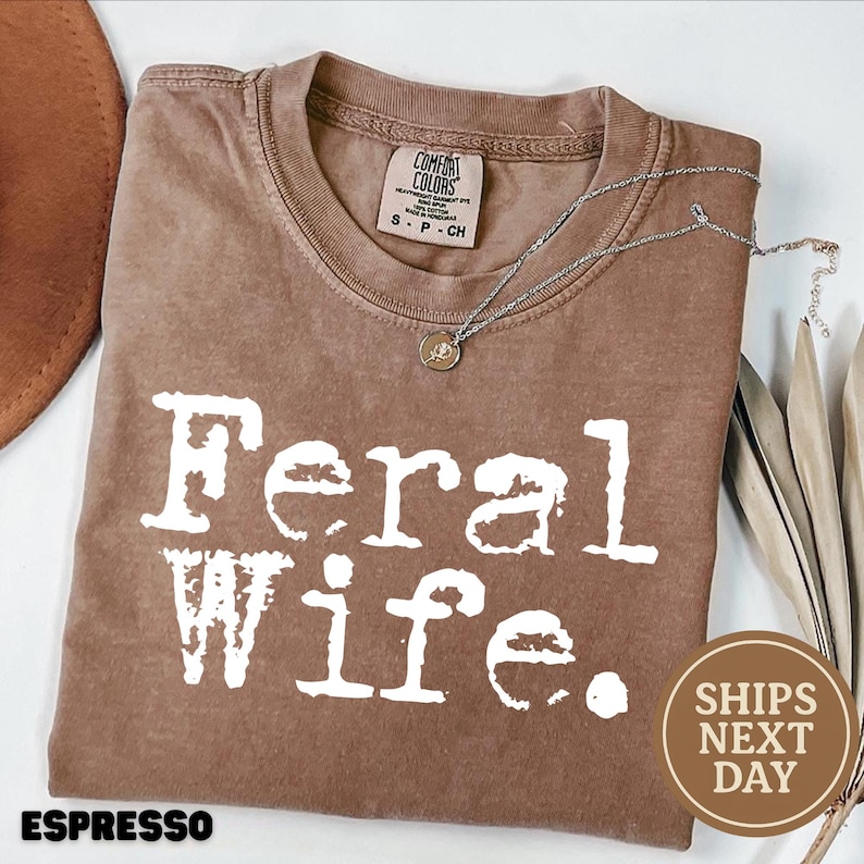 May include: A brown t-shirt with the text "Feral Wife." printed in white. The shirt has a distressed look and is made of a soft, comfortable fabric.