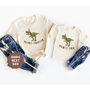 May include: Two cream-coloured t-shirts with dinosaur graphics and text. One shirt says "MAMA-SAURUS" and the other says "THREE-REX". Both shirts have a green dinosaur wearing a party hat. The shirts are paired with blue jeans.