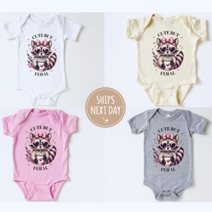 Pu&ograve; includere: Quattro body per neonati bianchi, crema, rosa e grigi, ciascuno con una grafica di procione con occhiali da sole a forma di cuore e la scritta "CUTE BUT FERAL". Il procione tiene in mano una tazza di caff&egrave;. L'immagine include una grafica "SHIPS NEXT DAY".