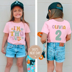 Może przedstawiać: R&oacute;żowa koszulka z napisem "I'm Two Cute" w kolorowych literach. Koszulkę nosi dziecko w czarnej czapce z daszkiem. Na plecach koszulki widnieje imię "Olivia" i liczba "2" w kolorowych literach.