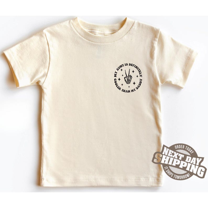 May include: A cream coloured t-shirt with a black circle graphic that says "My Aunt is Definitely Cooler Than My Daddy" with a skeleton hand making a peace sign.
