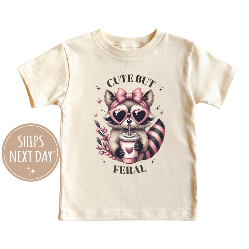Pu&ograve; includere: T-shirt color crema con un'illustrazione di procione. Il procione indossa occhiali da sole a forma di cuore e un fiocco, tenendo in mano una bevanda con un cuore. Il testo dice "CUTE BUT FERAL".