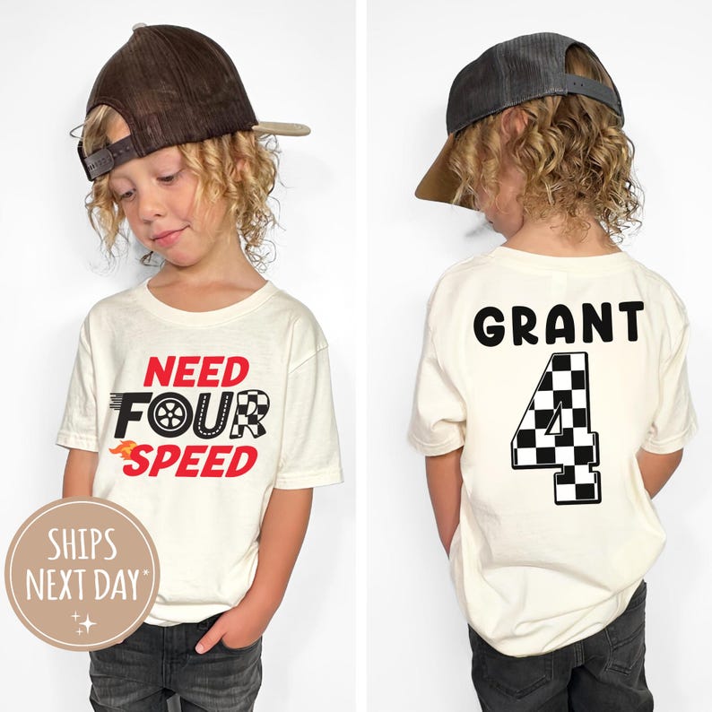 以下が含まれることがあります： 白地に「Need Four Speed」の文字が書かれたTシャツ。後ろには「Grant」という名前と、市松模様の旗のデザインが施された「4」の数字が書かれている。茶色のベースボールキャップをかぶった子供が着用している。