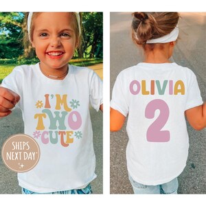 Może przedstawiać: Dwie białe koszulki z kolorowym tekstem w stylu retro. Jedna koszulka ma napis "I'm Two Cute" z kwiatami, a druga "Olivia 2".